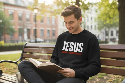 Mann sitzt auf einer Parkbank und liest, während er ein christliches Sweatshirt mit dem Schriftzug „Jesus – The Way, The Truth, The Life“ trägt